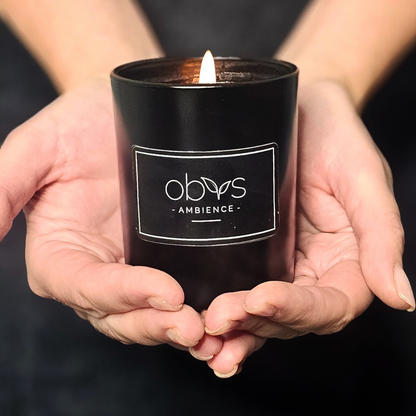 A matte black candle jar filled with organic essential oils, burning with a soft, warm flame. The minimalist design exudes elegance, while the gentle aroma from natural essential oils creates a calming, eco-friendly atmosphere. The candle sits on a wooden surface, with subtle shadows enhancing its luxurious, earthy vibe, perfect for relaxation or meditation