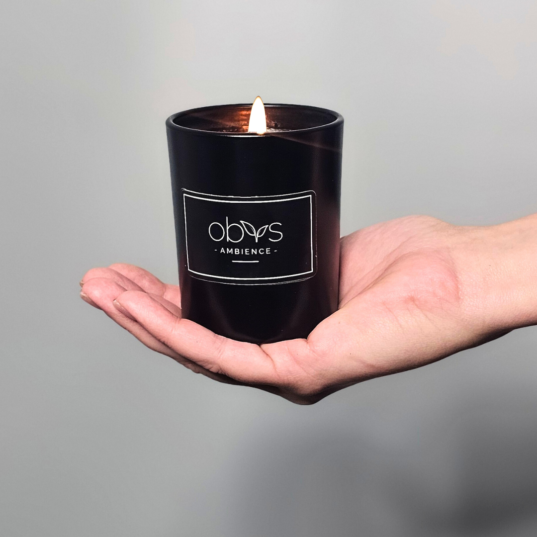 A matte black candle jar filled with organic essential oils, burning with a soft, warm flame. The minimalist design exudes elegance, while the gentle aroma from natural essential oils creates a calming, eco-friendly atmosphere. The candle sits on a wooden surface, with subtle shadows enhancing its luxurious, earthy vibe, perfect for relaxation or meditation