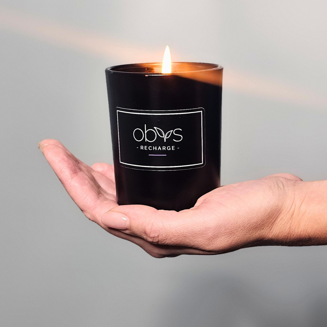 A matte black candle jar filled with organic essential oils, burning with a soft, warm flame. The minimalist design exudes elegance, while the gentle aroma from natural essential oils creates a calming, eco-friendly atmosphere. The candle sits on a wooden surface, with subtle shadows enhancing its luxurious, earthy vibe, perfect for relaxation or meditation