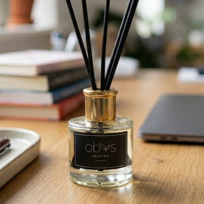 A diffuser bottle with black sticks on a wooden surface with books and a laptop in the background.