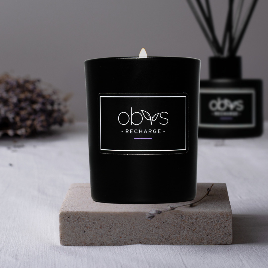 A matte black candle jar filled with organic essential oils, burning with a soft, warm flame. The minimalist design exudes elegance, while the gentle aroma from natural essential oils creates a calming, eco-friendly atmosphere. The candle sits on a wooden surface, with subtle shadows enhancing its luxurious, earthy vibe, perfect for relaxation or meditation