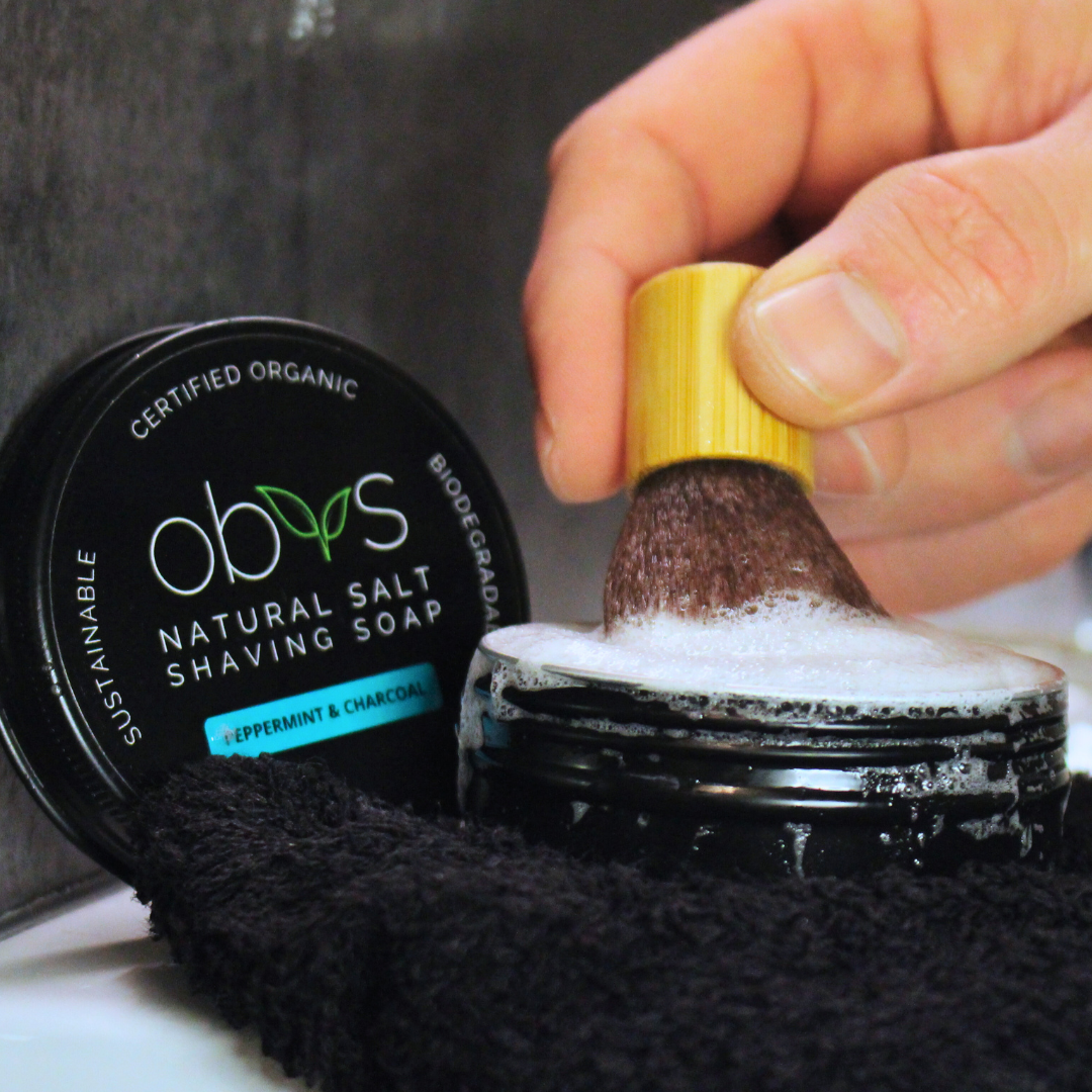 Hand using a shaving brush with a jar of Obv's Natural Salt Shaving Soap on a towel. Obvs Skincare New Shaving Soap Travel Kit with peppermint and charcoal shaving soap in travel tin and mini shaving brush