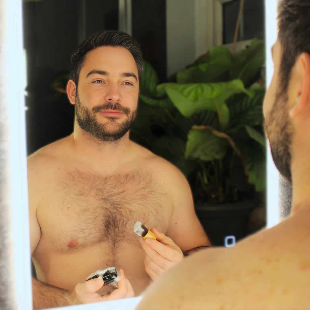 Man shaving his chest in front of a mirror with plants in the background Obvs Skincare New Shaving Soap Travel Kit with peppermint and charcoal shaving soap in travel tin and mini shaving brush