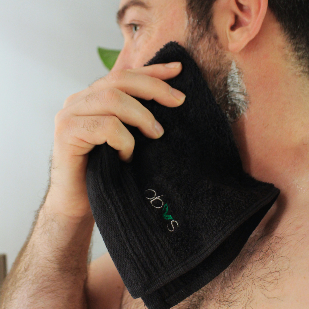Man using a black organic face cloth towel with an obvs logo on his face, against a neutral background